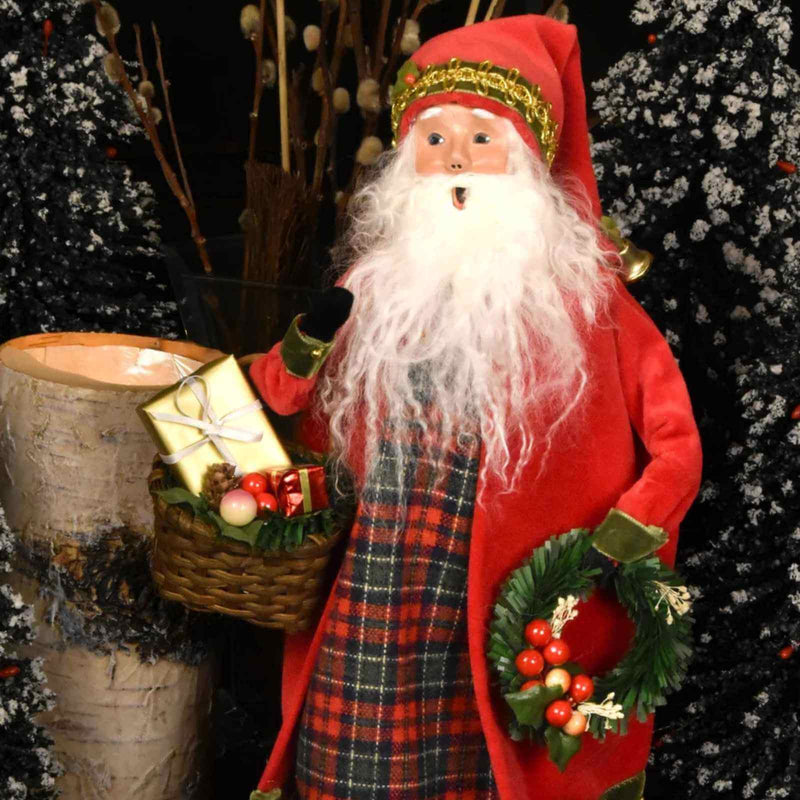A Santa Claus figurine wearing a red coat and hat with a long white beard, holding a basket of wrapped gifts and a green wreath with red berries, posed in front of dark decorated trees and a birch log.
