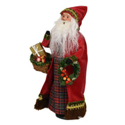 A Christmas Santa Claus figurine wearing a red velvet robe with gold trim, a red and black plaid skirt, and a long white beard, holding a gift basket and a green wreath with red berries, isolated on a white background.